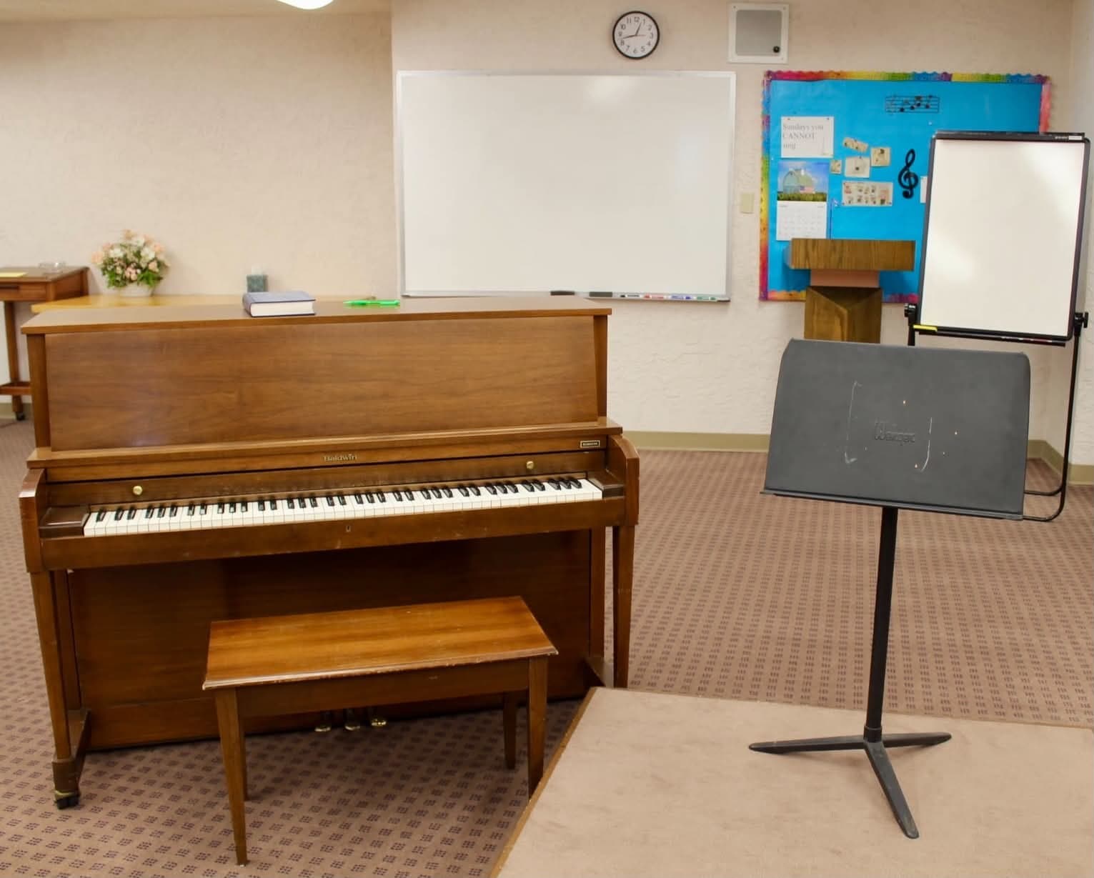 The choir room, with an upright Baldwin piano, a whiteboard and a music stand.