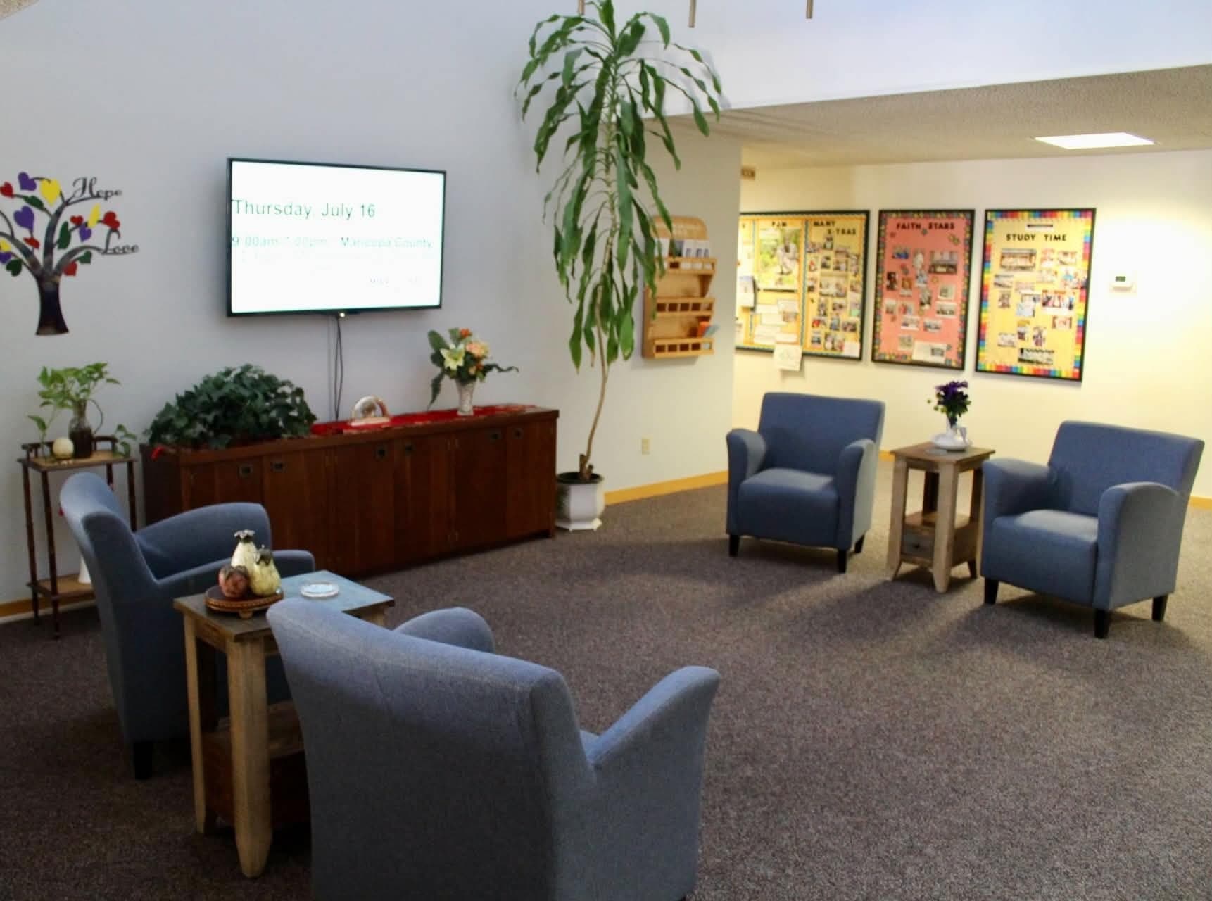 A carpeted waiting area with armchairs arranged around side tables and a large plant.